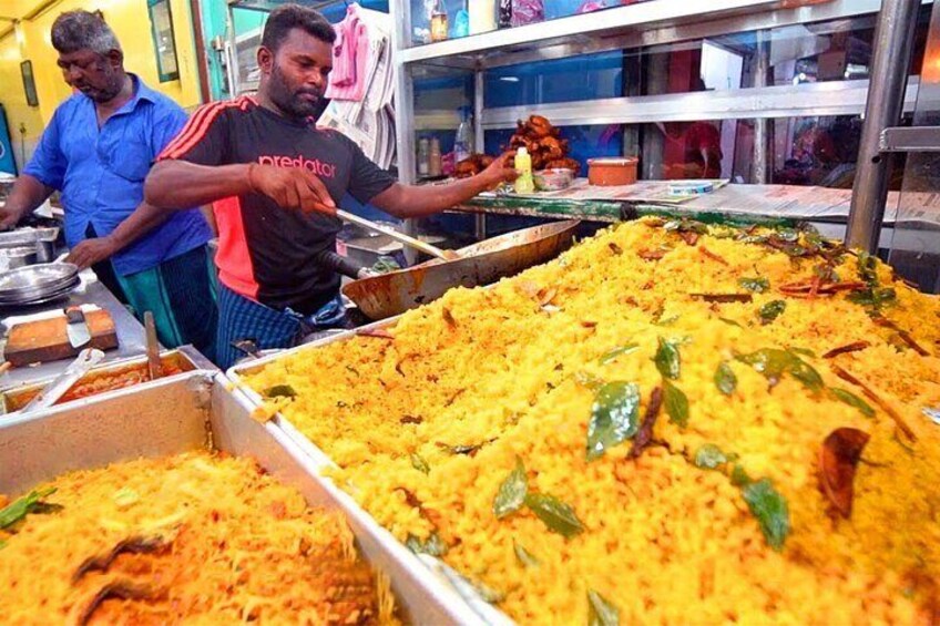 Un puesto de comida con grandes bandejas de arroz amarillo, una persona sirviendo y otra al fondo. 2