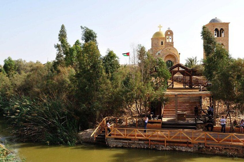Een houten constructie met een trap naar een platform, omgeven door groen en een rivier. Achtergrond toont een kerk met een klokkentoren en een vlag. 5