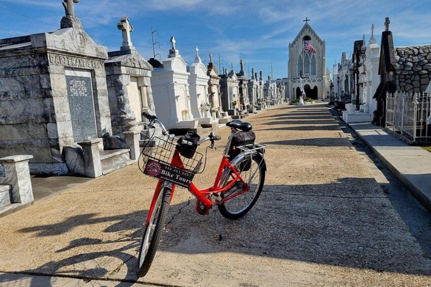 Una bicicleta roja estacionada en un sendero de grava en un cementerio con lápidas blancas y una iglesia al fondo. 2
