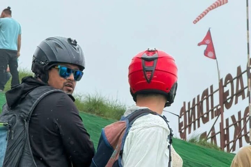 A person wearing a red helmet and a backpack, standing on a grassy slope with a sign in the background. 2