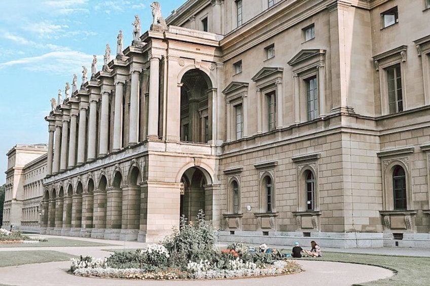 A large classical building with columns and statues, a central archway, and a landscaped garden with flowers. 5