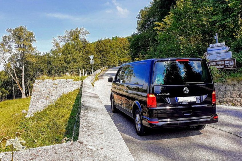 A black Volkswagen van parked on a road with a stone wall and greenery on one side. 3