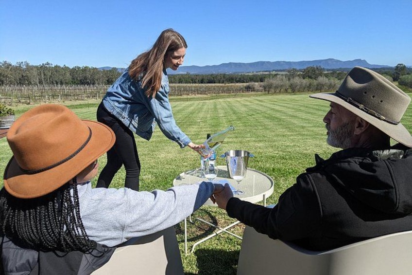 A group of people at an outdoor table with a clear view of a vineyard and mountains in the background. 2