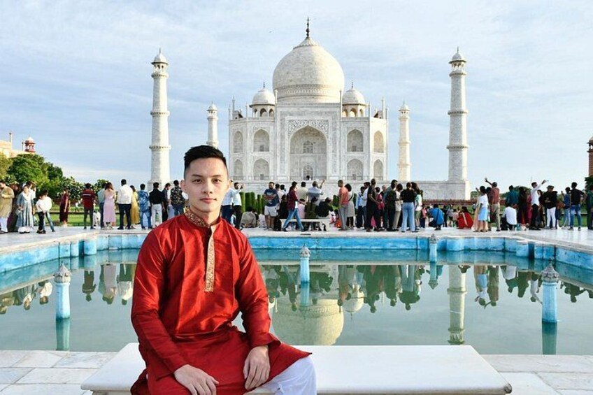 A person in a red traditional outfit sitting by a reflective pool with the Taj Mahal in the background. 3