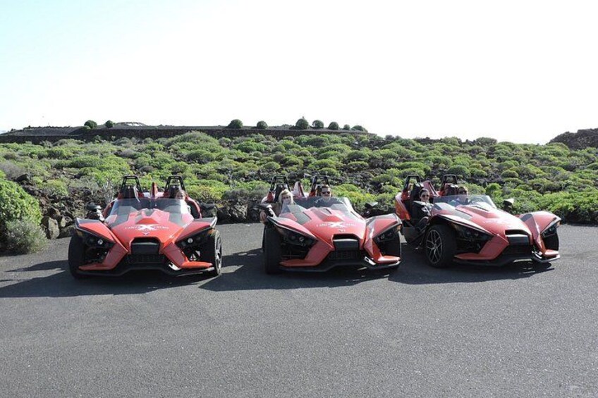 Three red sports cars parked on a paved surface with a hilly landscape in the background. 2