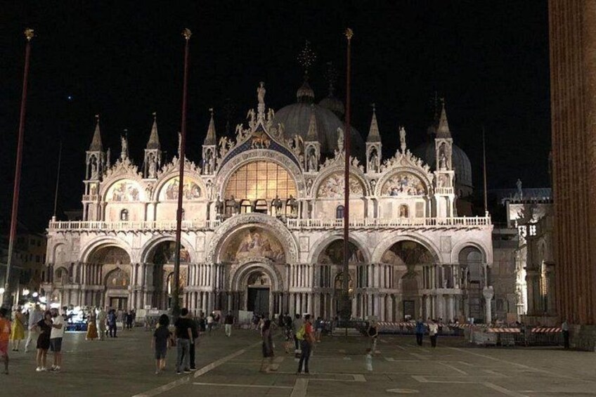 A grand, illuminated cathedral with multiple domes and spires, surrounded by a plaza with people walking. 1