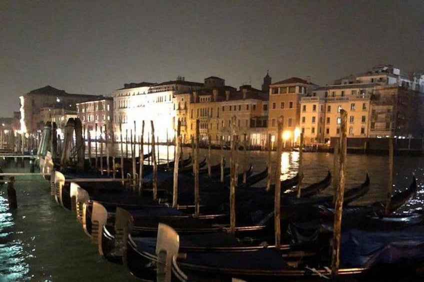 A nighttime view of a canal with gondolas docked along the shore, buildings lining the water, and a bridge in the background. 3