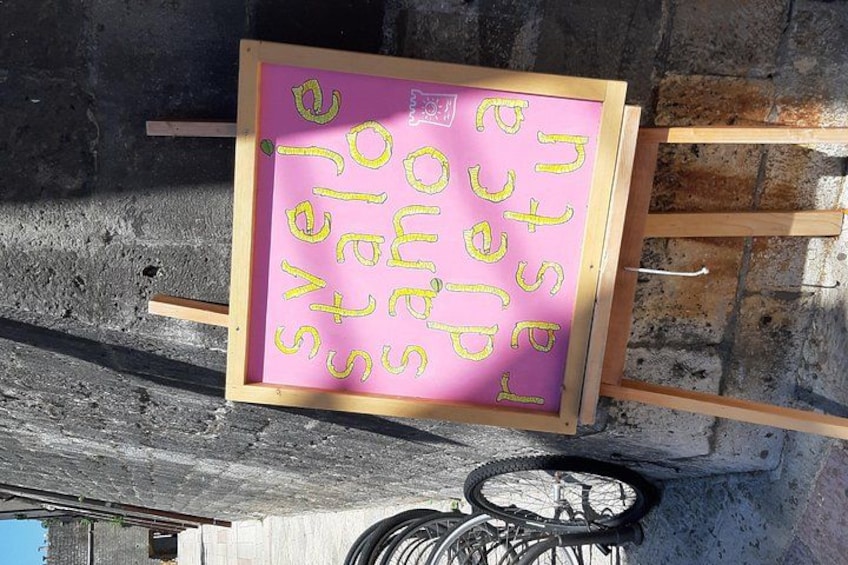 A pink sign with yellow text on a wooden frame, mounted on a wall with a bicycle wheel visible below. 5