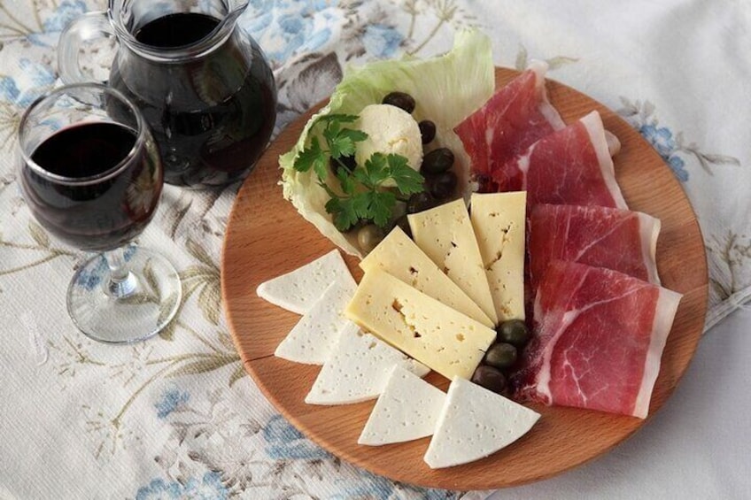 A wooden plate with cheese, prosciutto, olives, and a salad on a tablecloth. 3