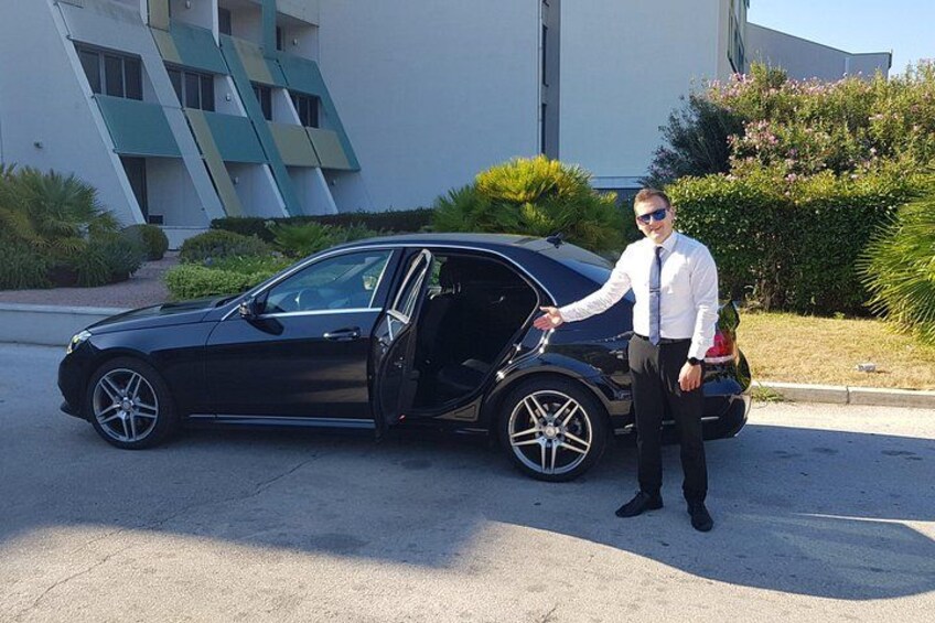 A man standing next to a black car with the door open, a building with a unique design, and some greenery in the background. 2