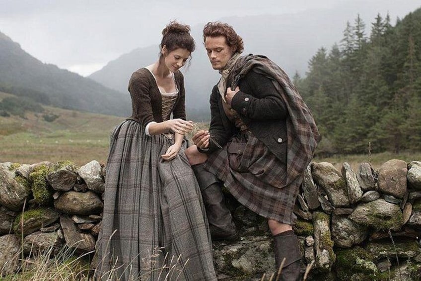 A person in a plaid coat sitting on a stone wall, another person in a dress standing beside them, and a mountainous landscape in the background. 2