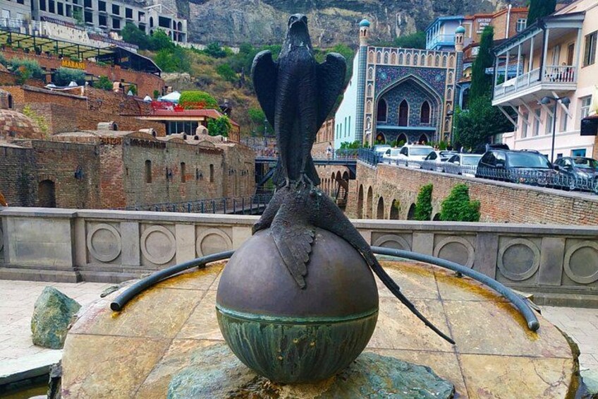 Una scultura in bronzo di una creatura alata su un piedistallo, con montagne sullo sfondo e architettura tradizionale. 2