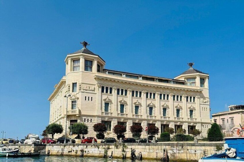 A large, historic building with a prominent dome and columns, situated by a waterfront with parked cars and boats. 3