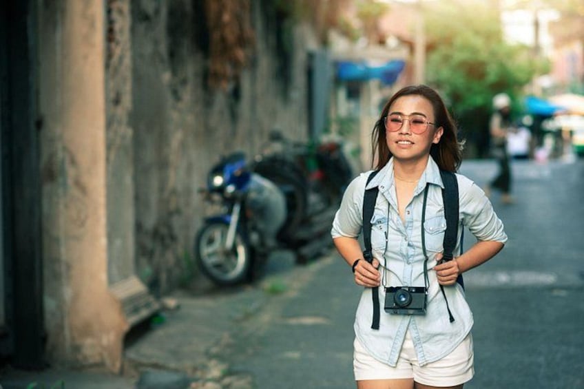 A person walking down a narrow street with a camera around their neck, a motorcycle parked on the side, and a building with a weathered facade. 2