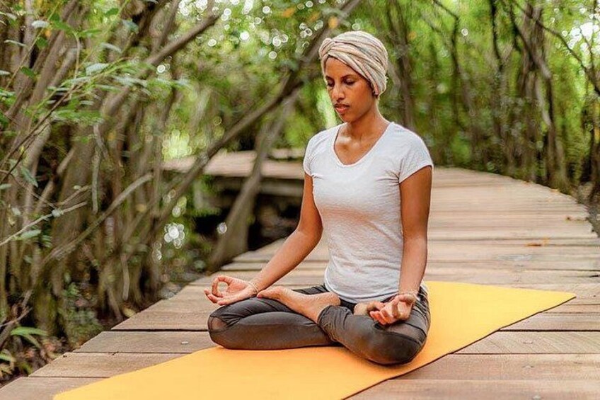 A person sitting cross-legged on a yoga mat in a forested area with a wooden bridge in the background. 3