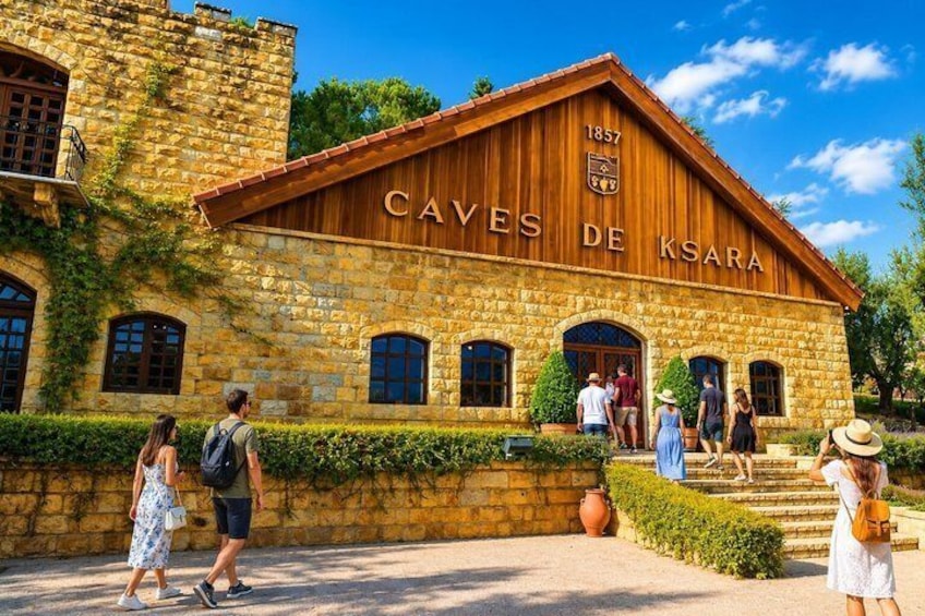 Un edificio storico chiamato 'Caves de Ksara', con mura in pietra e tetto in legno, immerso nel verde e circondato da visitatori. 5