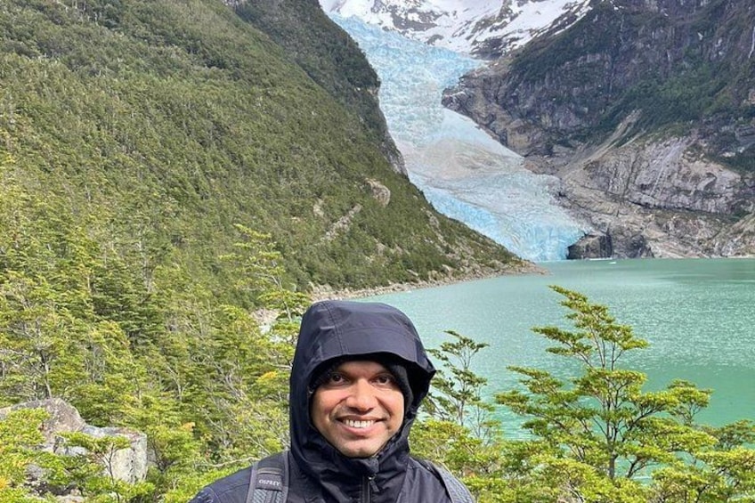 A glacier with a person in a black jacket and hood, a green lake, and a mountainous landscape. 2