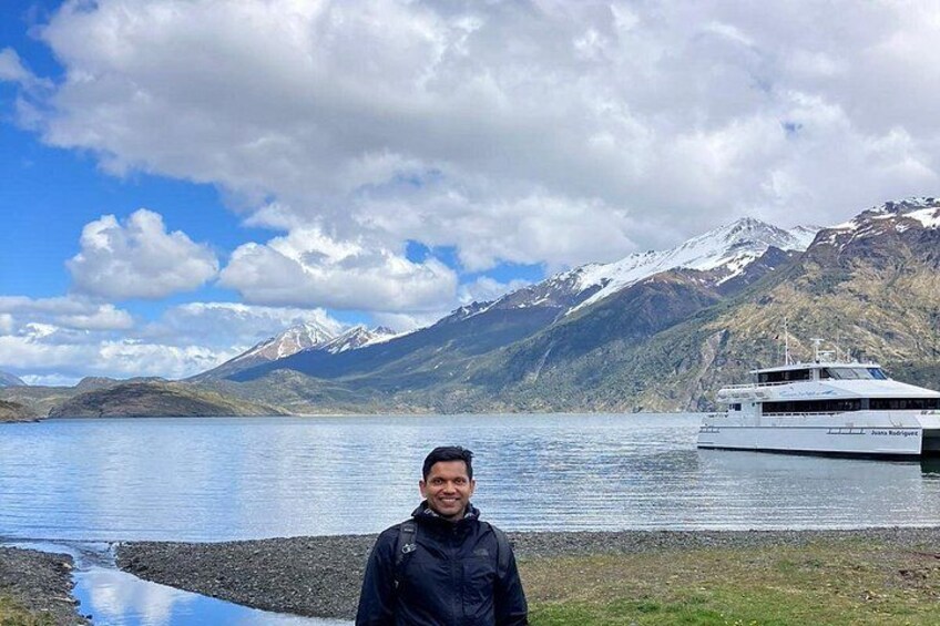 A person standing by a lake with mountains in the background and a boat on the water. 3