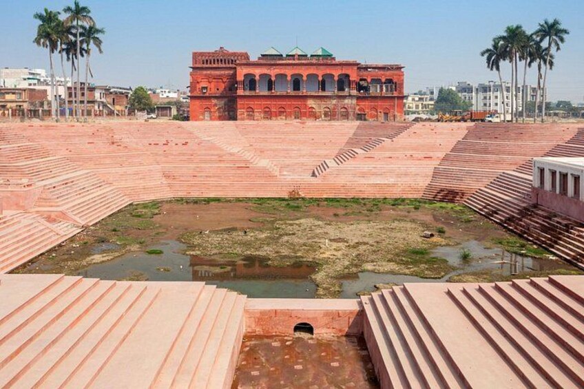 A large open area with tiered seating, a central water feature, and a prominent red building with arches in the background. 2