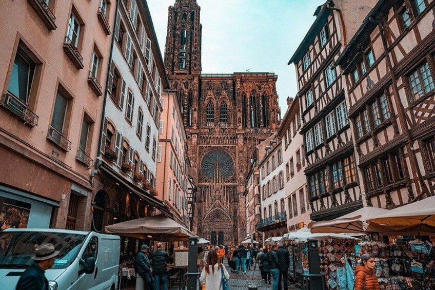 A bustling street with historic buildings, a prominent cathedral, and market stalls. 1