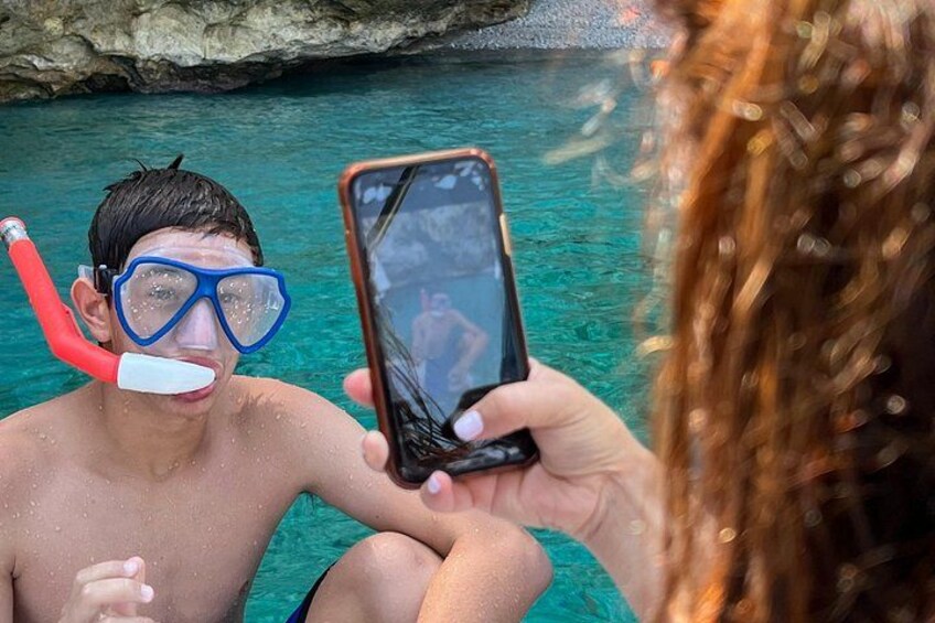 En person snorkler med en rød snorkel og blå dykkerbriller, og tager et selfie med en smartphone. 4