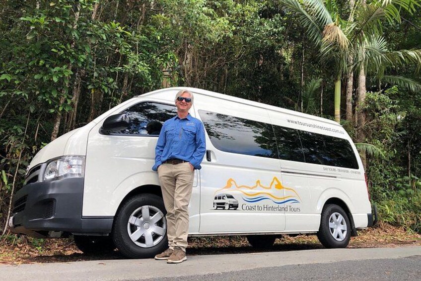 Un fourgon blanc arborant le logo « Coast to Hinterland Tours » est garé sur une route, avec un feuillage dense en arrière-plan. 1