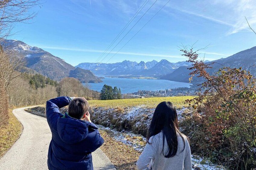Dos personas de pie en una carretera, contemplando un paisaje montañoso con un lago y picos nevados. 2
