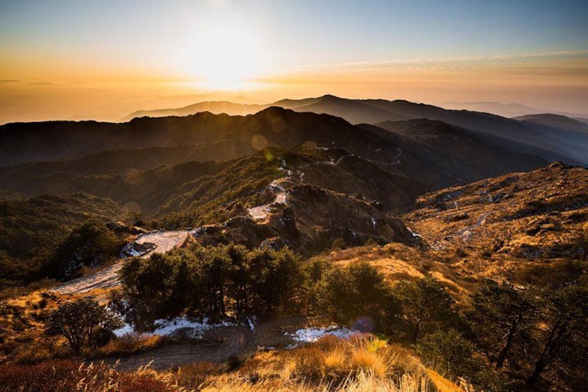 A mountainous landscape with a winding path, a small settlement, and a sunrise or sunset. 1