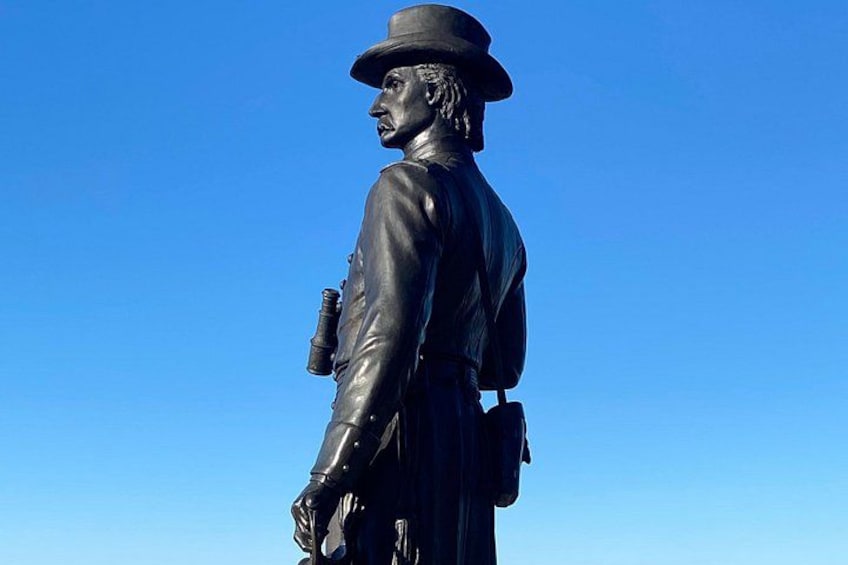 En staty av en man i kostym och hatt, som håller i en pistol, med en klarblå himmel i bakgrunden. 2