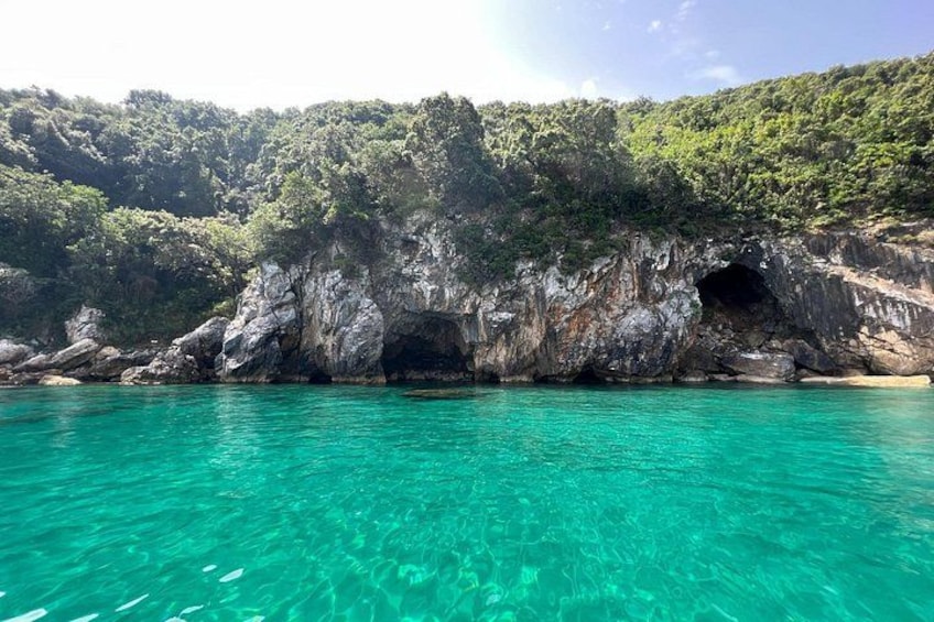 A coastal landscape with clear turquoise water, rocky cliffs, and dense green vegetation. 5