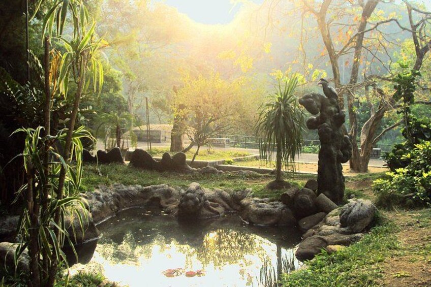 A pond surrounded by rocks and vegetation, with trees and a statue in the background. 5
