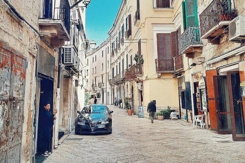 Une ruelle étroite pavée, bordée de bâtiments historiques, avec une voiture garée et une personne qui s’éloigne de la caméra. 1