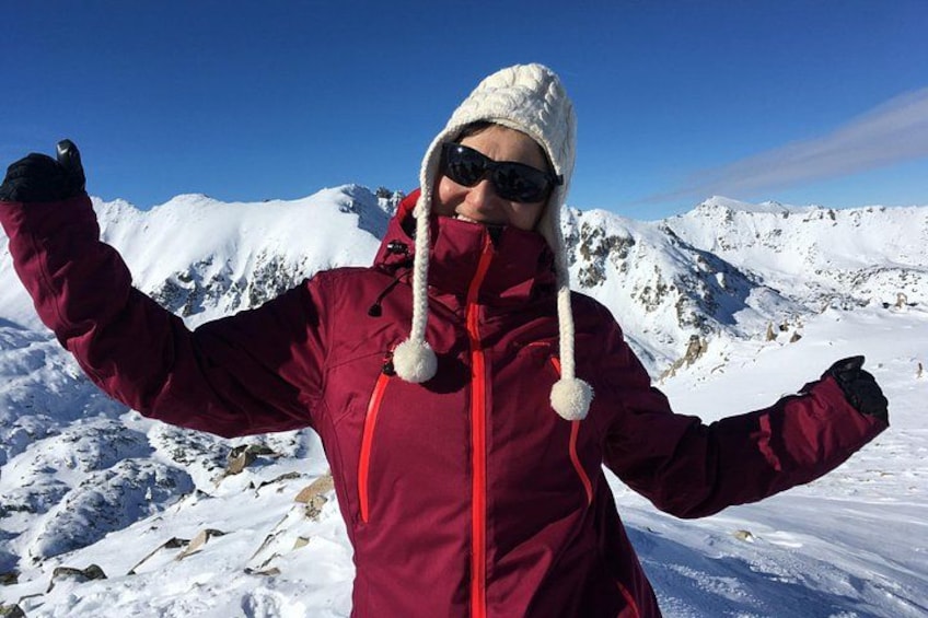 Une personne vêtue d’une veste rouge et d’un chapeau blanc, debout sur une montagne enneigée, sous un ciel bleu dégagé. 1
