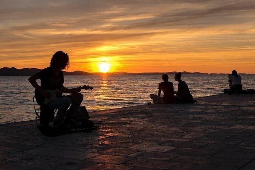 Un coucher de soleil sur l’eau, avec des personnes assises et une autre qui joue de la guitare. 2
