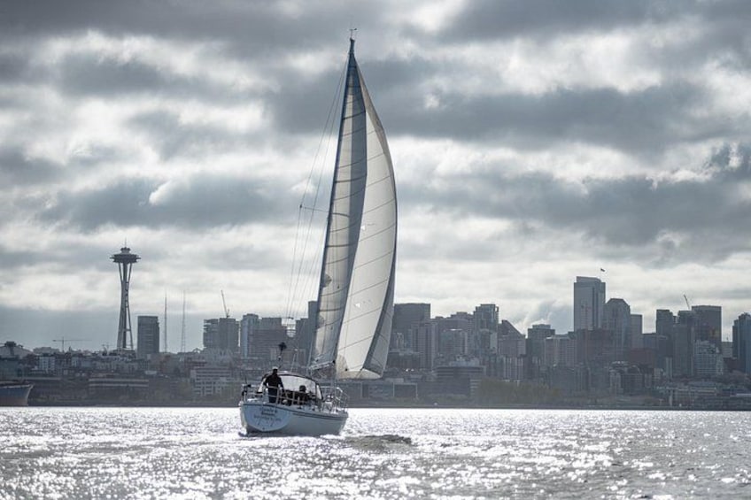 Un velero con una vela grande sobre una masa de agua, una silueta urbana con una torre destacada al fondo y un cielo nublado. 1