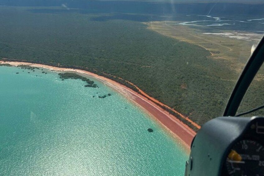 Vue depuis le cockpit d’un hélicoptère sur une zone côtière, avec une mer turquoise limpide, une étroite plage de sable et une forêt verdoyante et dense en arrière-plan. 2