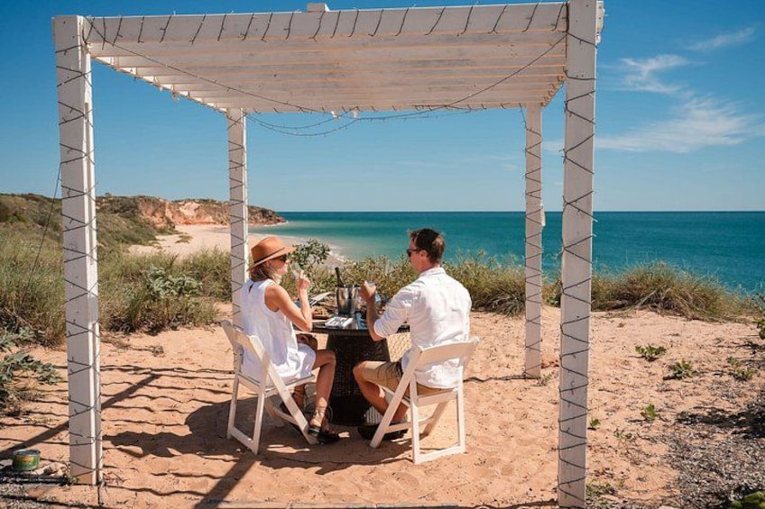A couple dining under a pergola on a beach with the ocean in the background. 2