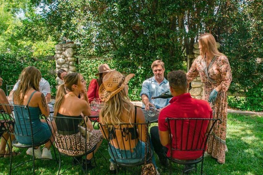 Un groupe de personnes est assis à une table en plein air, entouré de verdure et d’une structure en pierre. 2