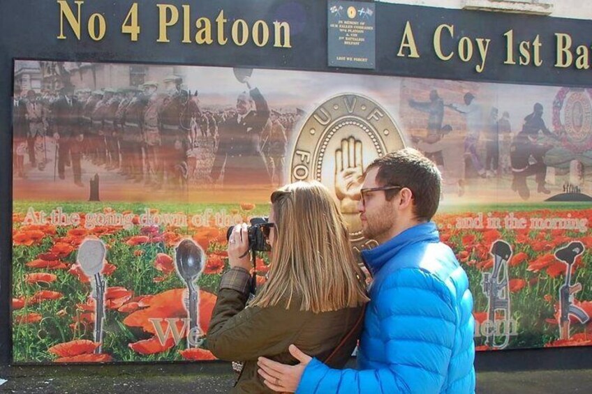 A couple taking a photo in front of a historical monument with a backdrop of red flowers and a large emblem. 2