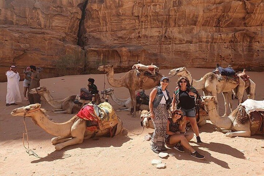 A group of people with camels in a desert setting with rocky cliffs in the background. 2