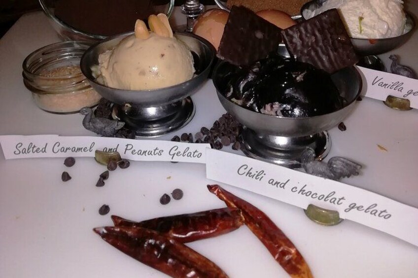 Two bowls of gelato, one labeled 'Salted Caramel and Peanuts Gelato' and the other 'Chili and Chocolate Gelato', with a chili pepper and chocolate pieces on the table. 5