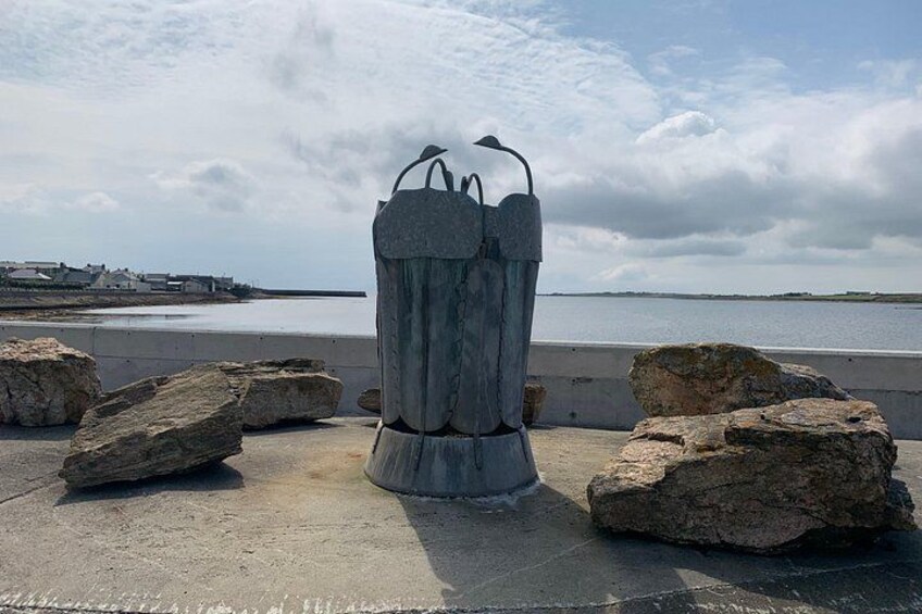 A metal sculpture with bird-like features on a concrete surface, surrounded by large rocks, with a body of water and a cloudy sky in the background. 2