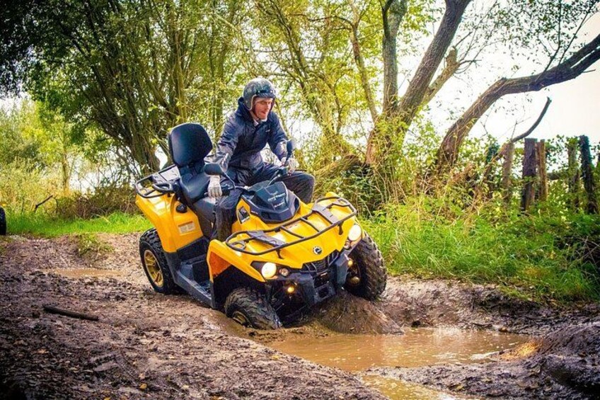 A person riding a yellow ATV on a muddy path with trees and a fence in the background. 3