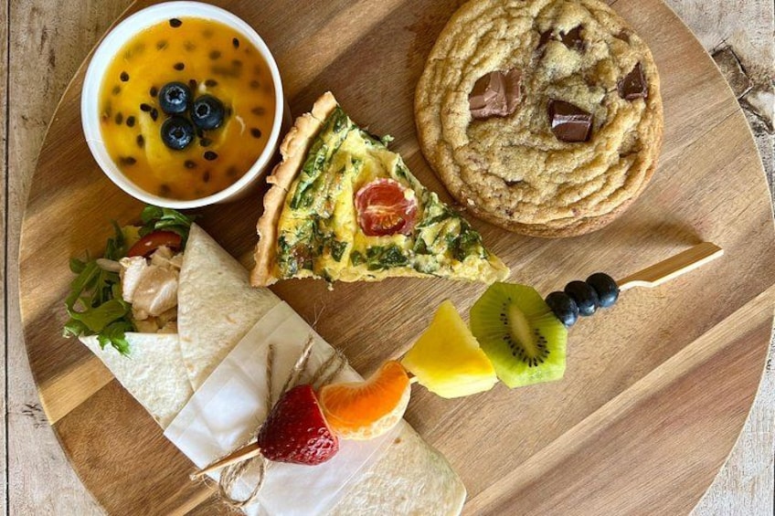 Une planche en bois sur laquelle on a disposé une part de quiche, un biscuit, un bol de soupe et une brochette de fruits. 2