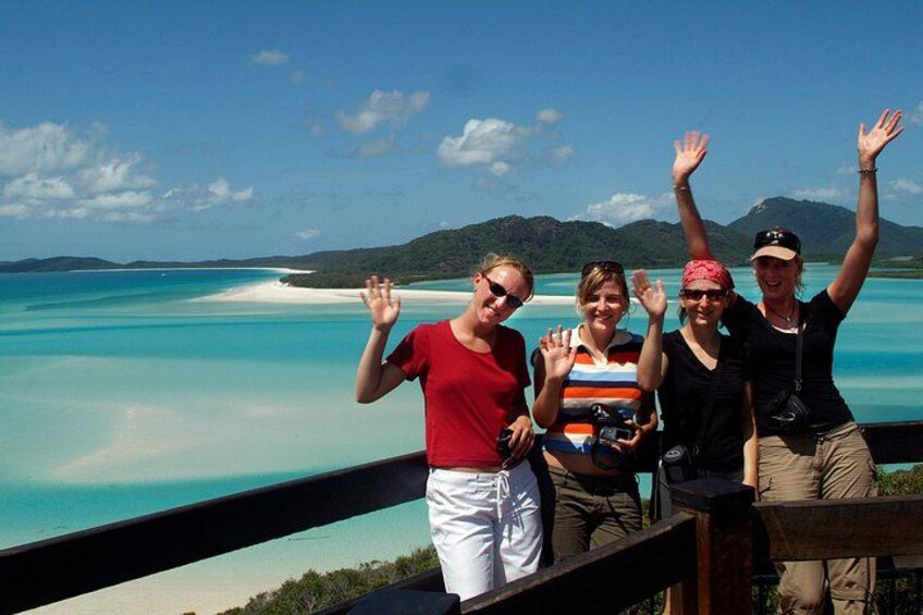 Un groupe de personnes se trouve sur une terrasse surplombant une mer turquoise et une plage de sable blanc, avec des montagnes en arrière-plan. 4