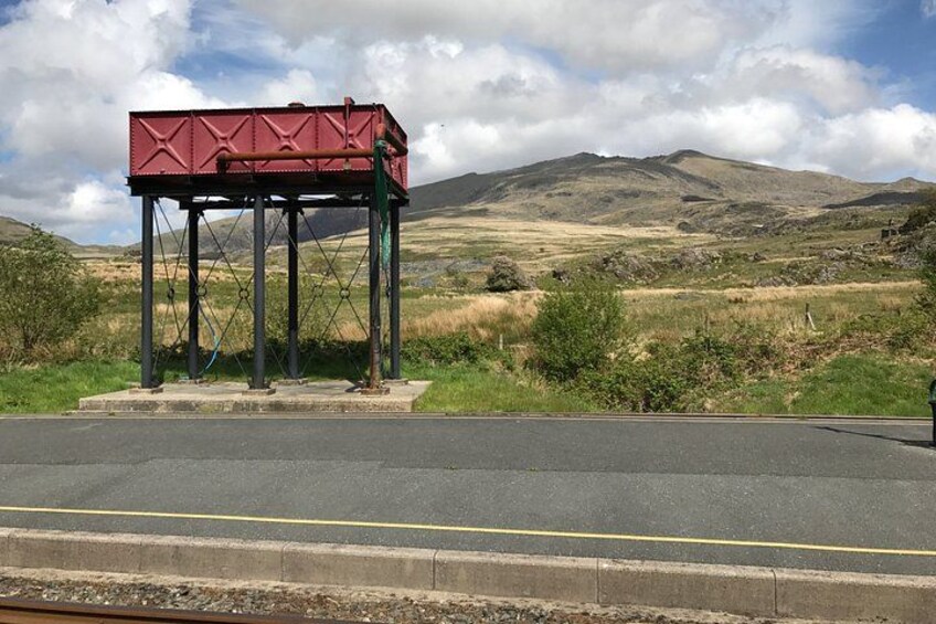 Une structure métallique rouge, montée sur des piliers, se dresse au bord d’une route, avec un paysage vallonné en arrière-plan. 3