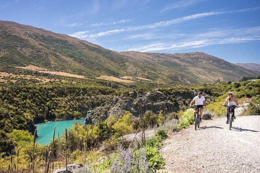 Due ciclisti su un sentiero sterrato, con un lago turchese e colline verdi sullo sfondo. 4