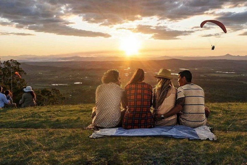 A group of people sitting on a blanket, watching a sunset with a parachute in the sky. 1