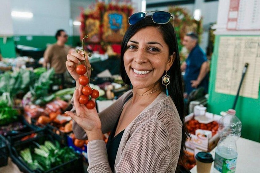 A person holding a bunch of tomatoes at a market with various vegetables and produce in the background. 2