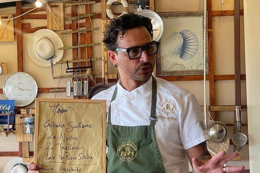 A person holding a menu with handwritten text, wearing a chef's apron, and standing in a kitchen with wooden walls and various kitchen utensils. 2